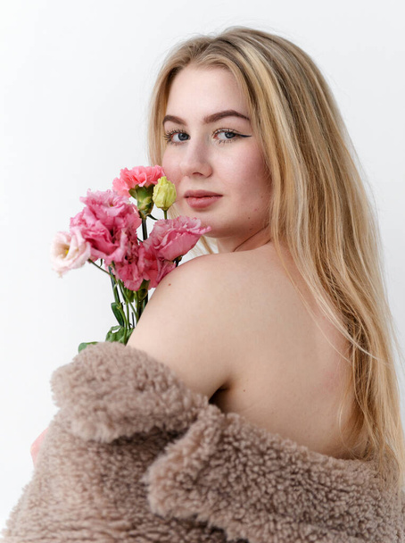 beautiful white young blonde woman in brown furry coat holds a bouquet of pink flowers and looks directly from her back on a white background - Фото, зображення