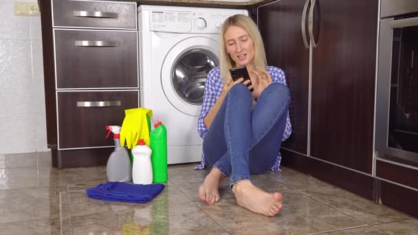 blondýny v džínách a košili sedí na podlaze v kuchyni v blízkosti pračky - Záběry, video