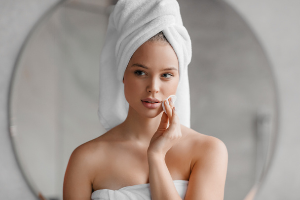 Rutyna piękna. Ładna młoda kobieta czyści twarz podkładką bawełnianą w domu, stojąc z ręcznikiem na głowie po kąpieli - Zdjęcie, obraz