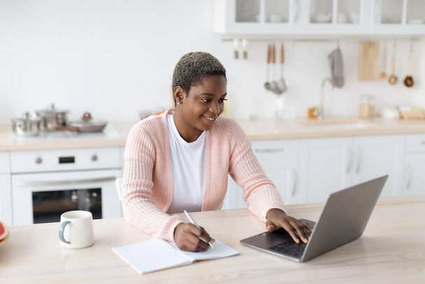 Sorrindo jovem afro-americana em casual trabalhando no laptop no interior da cozinha minimalista - Foto, Imagem