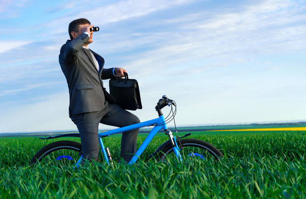 business concept - A businessman rides a bicycle on a green grass field, looks into the distance through binoculars, dressed in a business suit, he has a briefcase and documents - Foto, Imagem