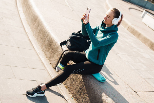 Young smiling sportswoman in headphones with prosthesis sitting on a fitness mat using mobile phone outdoors - Foto, immagini