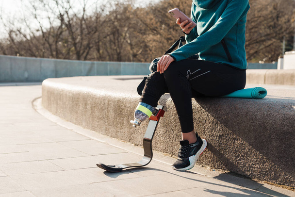 Young sportswoman with prosthesis using cellphone while working out outdoors - Foto, Bild