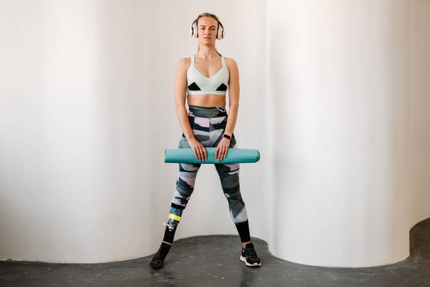 Young sportswoman with prosthesis posing with fitness mat indoors - Foto, Imagen