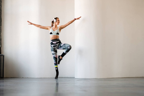 Young sportswoman with prosthesis listening music and dancing indoors - 写真・画像