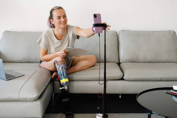 Smiling young white disabled woman blogger with prostetic leg sitting on a couch in front of a camera stand making a video at home - Fotoğraf, Görsel