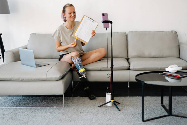 Smiling young white disabled woman blogger with prostetic leg sitting on a couch in front of a camera stand making a video at home making tutorial - Fotoğraf, Görsel