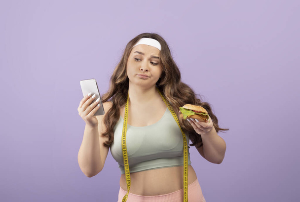 Insoddisfatto triste europeo giovane plus size femminile in abbigliamento sportivo con metro nastro tenere hamburger guardare il telefono - Foto, immagini
