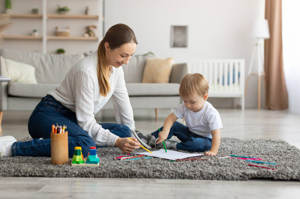 Ocio con bebé. Madre feliz e hijo pequeño dibujo con lápices de colores, relajarse juntos en el suelo en la sala de estar - Foto, Imagen
