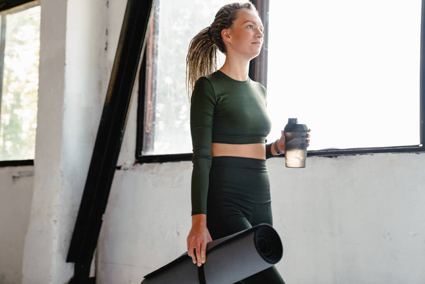 Young sportswoman holding fitness mat and water bottle indoors - 写真・画像