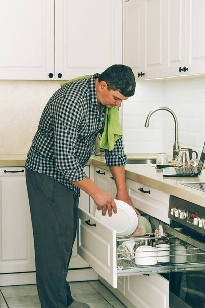 Um homem limpa pratos limpos com uma toalha e coloca-los para secar no armário da cozinha, tarefas domésticas diárias - Foto, Imagem