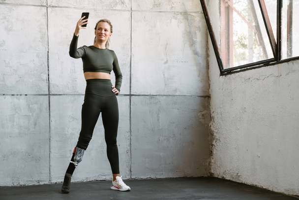 Young sportswoman with prosthesis taking selfie on cellphone indoors - Фото, зображення