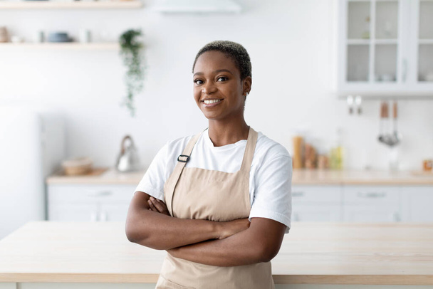 Fröhliche afrikanisch-amerikanische Amerikanerin in Schürze mit verschränkten Armen blickt in die Kamera moderner minimalistischer Küche - Foto, Bild