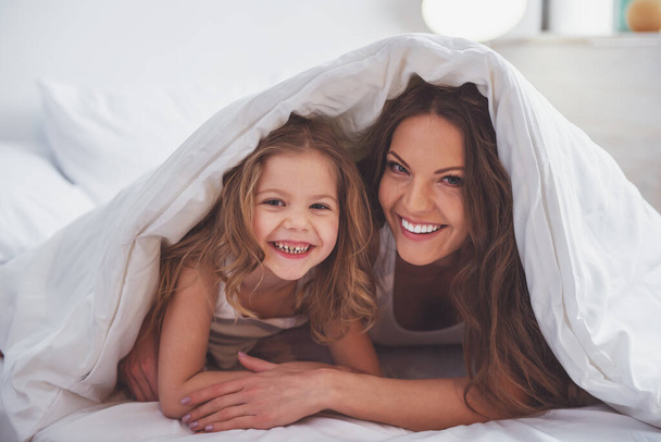 Słodkie dziewczynki i jej piękne młode matki są patrząc na kamery i uśmiechając się leżąc w łóżku pod kocem - Zdjęcie, obraz