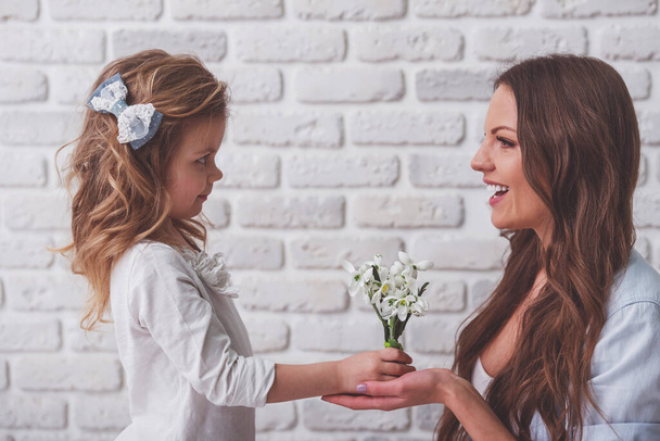 Aranyos kis lány ad virágok gyönyörű fiatal anya, fehér falhoz - Fotó, kép