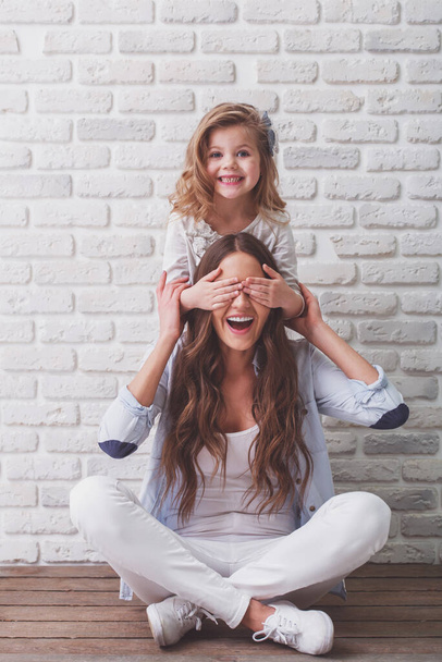 Young mother and her little daughter are looking at camera and smiling, sitting against white brick wall. Girl is closing her mother eyes - Photo, Image