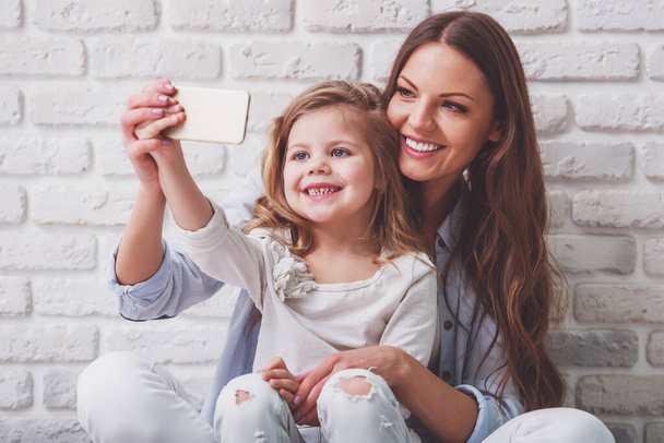 Kaunis nuori äiti tekee selfie hänen söpö pieni tytär älypuhelimella ja hymyillen, vastaan valkoinen tiili seinään
 - Valokuva, kuva
