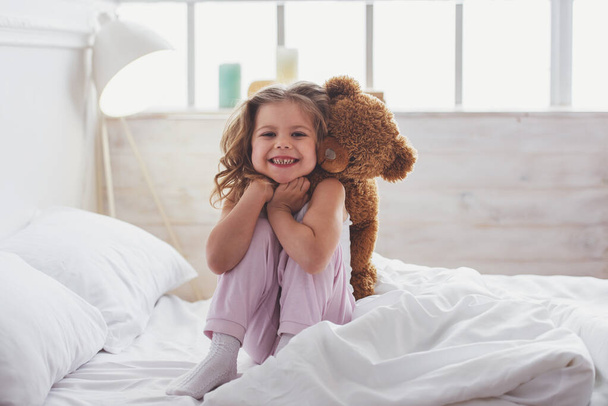 Sweet little girl is hugging a teddy bear, looking at camera and smiling while sitting on her bed at home - Photo, Image