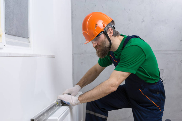 Un fontanero masculino instala un radiador en el sistema de calefacción de un apartamento. Un tipo con un mono y una llave de gas.. - Foto, imagen