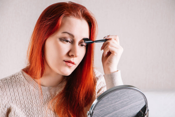 Mujer joven con el pelo rojo se aplica maquillaje. Lady se mira en el espejo y usa cosméticos.. - Foto, Imagen