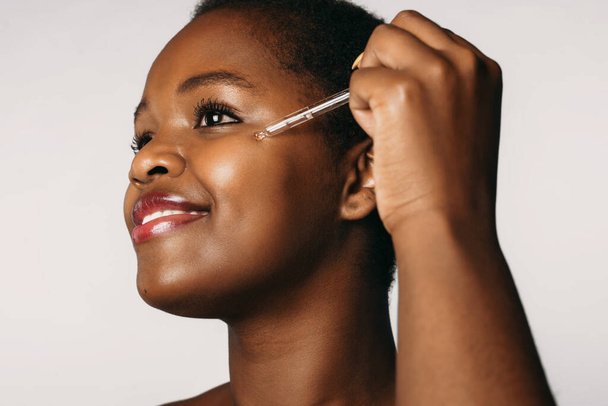 アフリカ系アメリカ人の女性が白い背景に隔離された彼女の顔に血清コラーゲン保湿剤をドロップします。美容整形手術。美顔. - 写真・画像