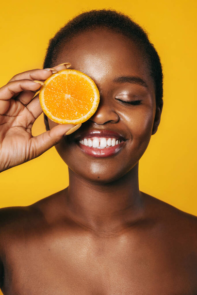 Retrato de close-up de uma mulher negra cobrindo o olho com uma fatia de laranja sorrindo com os olhos fechados isolados sobre o fundo laranja. Tratamento facial. Jovens lindos - Foto, Imagem
