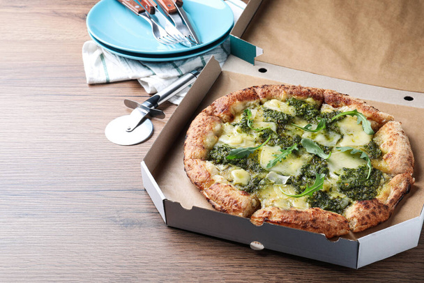 Delicious pizza with pesto, cheese and arugula in cardboard box served on wooden table - Φωτογραφία, εικόνα