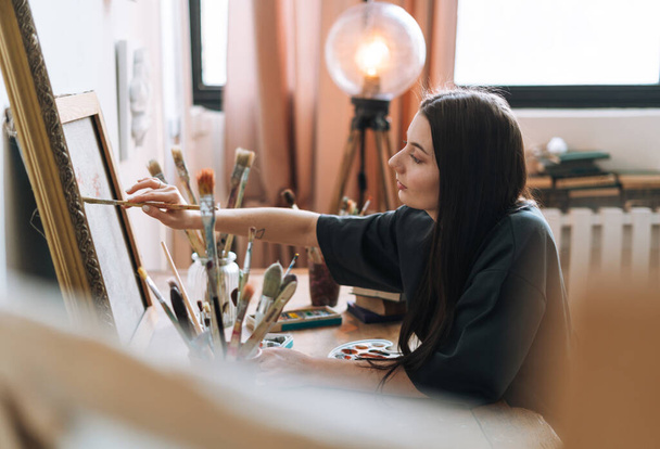 Koyu renk uzun saçlı genç kız ressam resim stüdyosunda resim çiziyor. - Fotoğraf, Görsel
