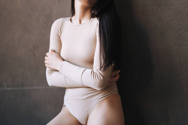 Young slim woman with dark long hair embracing body in beige color bodysuit against dark wall studio background, self loving girl - Φωτογραφία, εικόνα