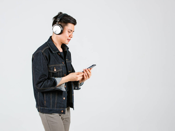 Portrait studio shot of Asian happy handsome male hipster model wearing casual street denim jeans jacket and headphone standing holding smartphone listening to music songs playlist on gray background. - Fotoğraf, Görsel