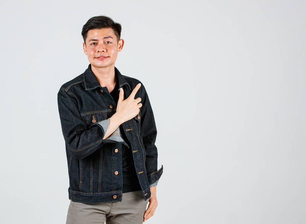 Studio strzał Azji młody przystojny mężczyzna hipster model noszenie casual ulicy dżinsy kurtka stoi trzymając się za ręce wskazując dwa palce wskazujące bok prezentując produkt reklamowy na szarym tle. - Zdjęcie, obraz
