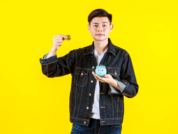 アジアの若い裕福な男のヒップスターモデルのポートレートスタジオショットカジュアルなストリートデニムジーンズジャケットを身に着けているカメラで見ると、黄色の背景に黄金のコインと貯金箱を示す. - 写真・画像