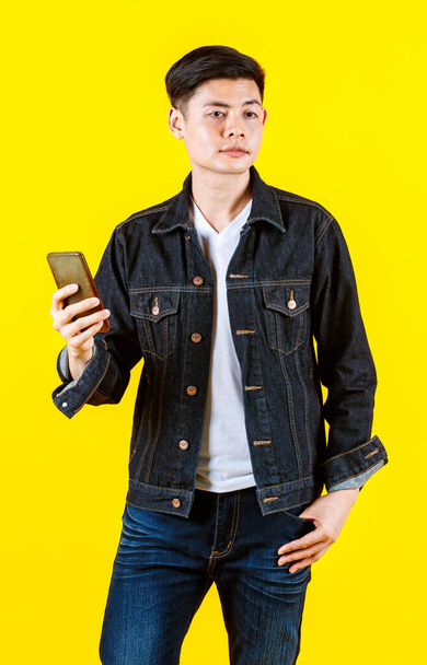 Muotokuva studio laukaus Aasian nuori rikas varakas mies hipster malli yllään rento katu denim farkut takki katso kameraa tilalla älypuhelin keltaisella taustalla. - Valokuva, kuva