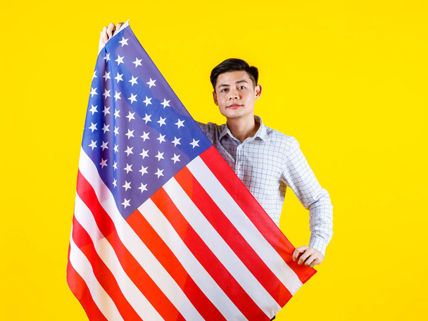 Portret studio shot van millennial aziatische jonge knappe mannelijke Amerikaanse burger zakenman model in casual outfit staande met grote Verenigde Staten van Amerika USA nationale vlag vel op gele achtergrond. - Foto, afbeelding