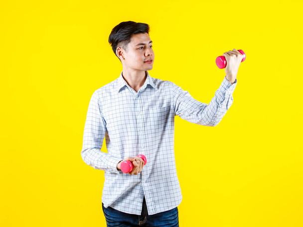 Portrait-Studioaufnahme von Millennial Asian junge hübsche männliche sportliche Athlet Modell in lässigem Outfit stehend Blick in die Kamera mit zwei kleinen rosafarbenen Hanteln heben Übungen auf gelbem Hintergrund. - Foto, Bild
