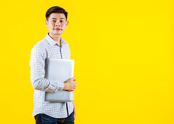 portret studio strzał z tysiąclecia Azji młody mężczyzna biznesmen w casual strój stojący uśmiechnięty gospodarstwa laptopa na żółtym tle - Zdjęcie, obraz