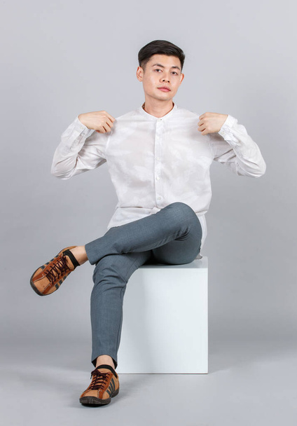 Estudio toma de millennial asiático guapo joven modelo masculino en casual traje sentado en blanco cuadrado taburete mirar cámara en gris fondo - Foto, imagen