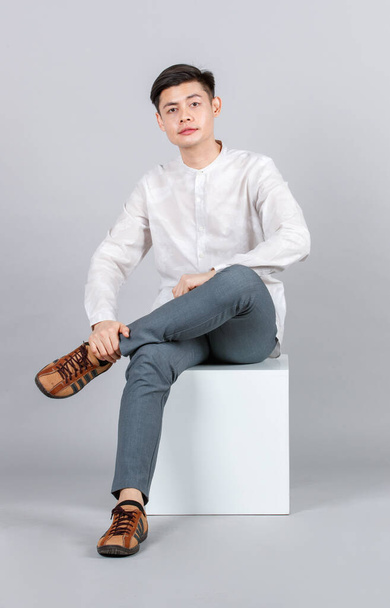 Studio shot of millennial Asian handsome young male model in casual outfit sitting on white square stool look at camera on gray background - Photo, image