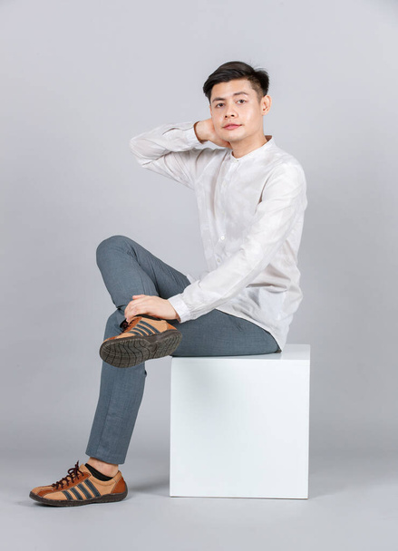 Studio shot of millennial Asian handsome young male model in casual outfit sitting on white square stool look at camera on gray background - Foto, immagini