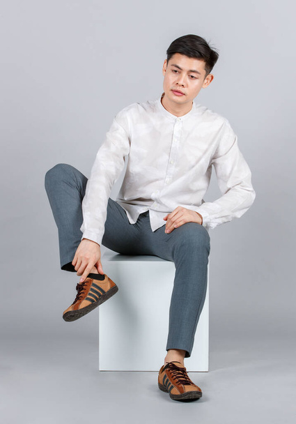 Studio shot of millennial Asian handsome young male model in casual outfit sitting on white square stool look at camera on gray background - Φωτογραφία, εικόνα