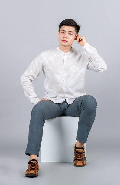 Estudio toma de millennial asiático guapo joven modelo masculino en casual traje sentado en blanco cuadrado taburete mirar cámara en gris fondo - Foto, Imagen