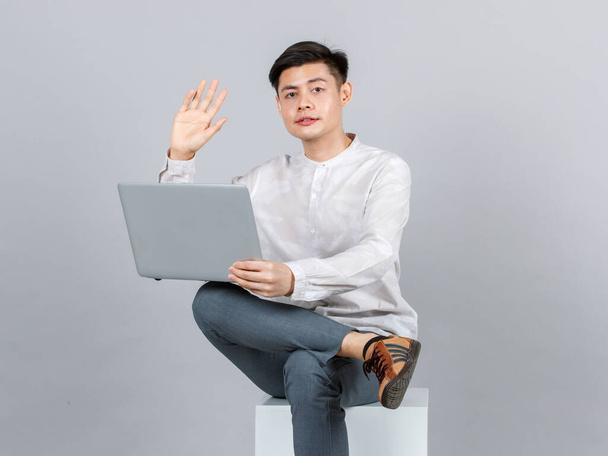 Στούντιο πλάνο της χιλιετίας Ασίας όμορφος νεαρός άνδρας μοντέλο σε casual στολή κάθεται σε λευκό τετράγωνο σκαμνί δείχνει εντάξει σήμα σήμα σήμα χέρι σε βιντεοκλήση με φορητό υπολογιστή σε γκρι φόντο. - Φωτογραφία, εικόνα