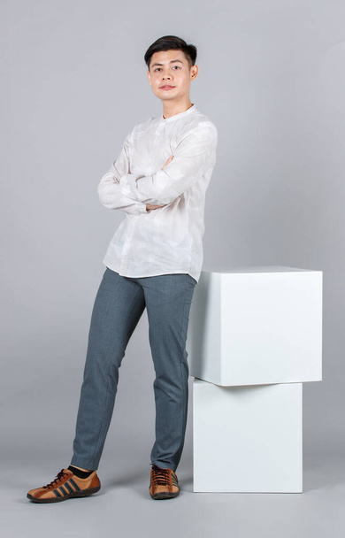 Portrait Studioaufnahme Millennial Asiatisch glücklich gutaussehendes männliches Modell in lässigem Langarmshirt-Outfit und Lederturnschuhen sitzend posierend auf weißem quadratischen Hocker lächelnd in die Kamera auf grauem Hintergrund. - Foto, Bild