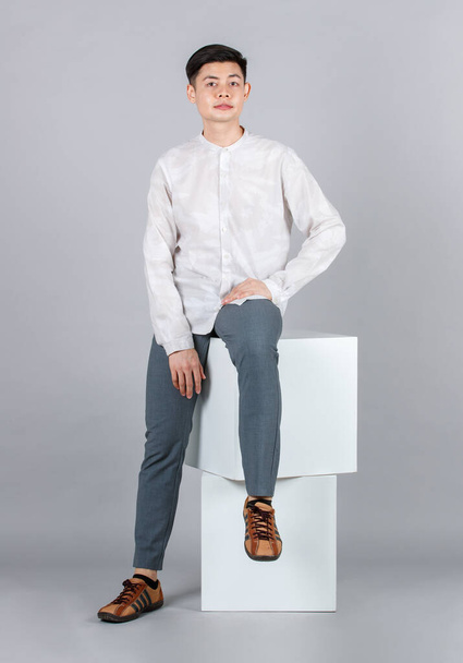 Portrait studio shot millennial Asian happy handsome male model in casual long sleeve shirt outfit and leather sneakers sitting posing on white square stool smiling look at camera on gray background. - Photo, Image
