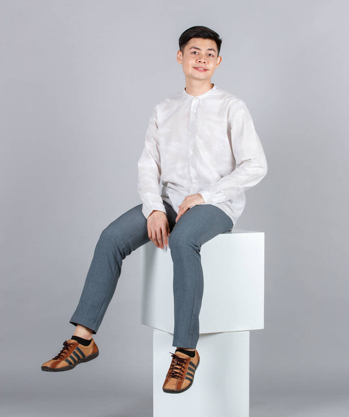Portrait Studioaufnahme Millennial Asiatisch glücklich gutaussehendes männliches Modell in lässigem Langarmshirt-Outfit und Lederturnschuhen sitzend posierend auf weißem quadratischen Hocker lächelnd in die Kamera auf grauem Hintergrund. - Foto, Bild