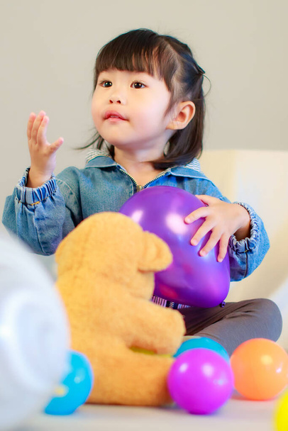 Studio shot of little cute preschooler girl daughter in casual street wears jeans jacket and sneakers sitting smiling teddy bear doll on floor playing with colorful plastic balls on gray background - Фото, зображення