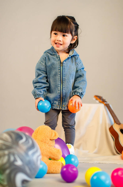 スタジオフルボディショットの小さなかわいい幼稚園未就学児の娘でカジュアル通り身に着けているジーンズジャケットとスニーカー立っているポーズ色鮮やかなプラスチックボールおもちゃで灰色の背景. - 写真・画像