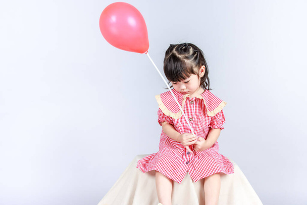 Στούντιο κοντινό πλάνο του μικρού χαριτωμένο προγυμναστή κοριτσάκι κόρη σε κόκκινο μακρύ φόρεμα κάθεται μόνη της στο περίπτερο κάλυψη με ύφασμα κρατώντας ήλιο αέρα κόμμα μπαλόνι ραβδί στο χέρι χαμογελώντας σε γκρι φόντο. - Φωτογραφία, εικόνα