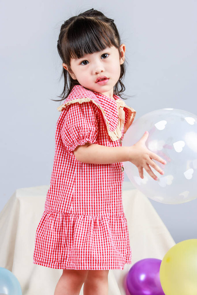 Portrait studio shot of little cute kindergarten preschooler kid girl daughter in red long dress standing smiling alone on floor playing with colorful helium air party balloons on gray background - Foto, Bild