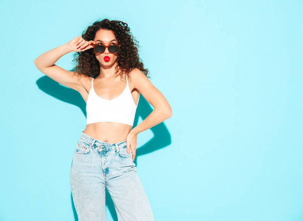 Young beautiful smiling female in trendy summer  hipster clothes. carefree woman posing near blue wall in studio. Positive brunette model with curls hairstyle. Cheerful and happy. In sunglasses - Zdjęcie, obraz